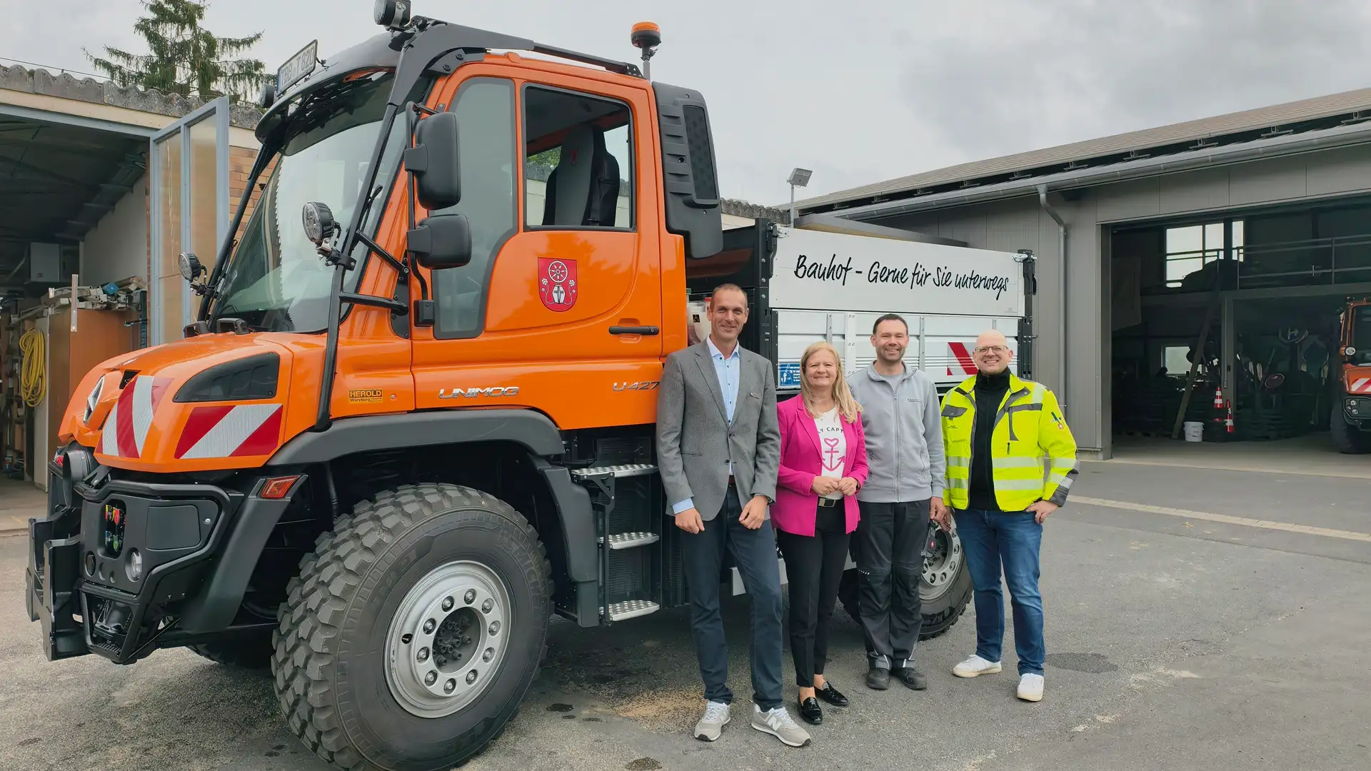 Ein Mercedes-Benz Unimog steht vor einem Gebäude im Bauhof; davor sind mehrere Personen für ein Gruppenfoto angeordnet.